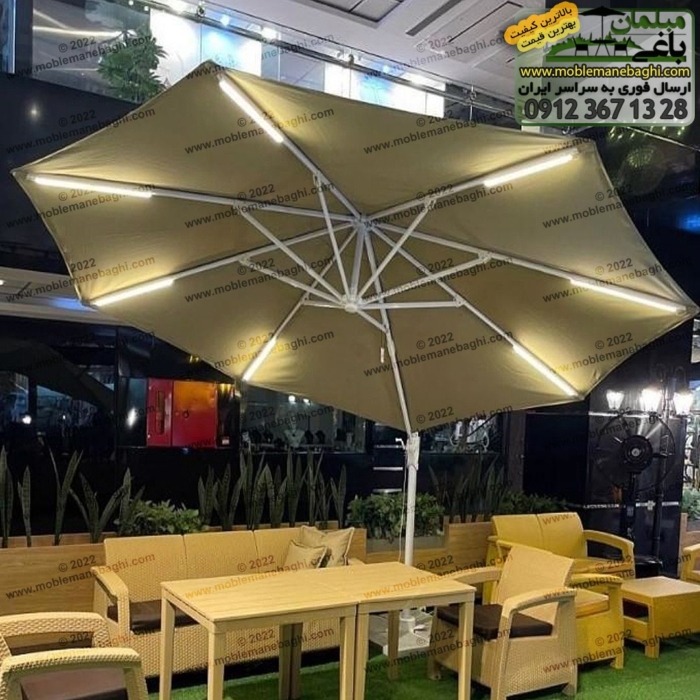 چتر باغی پایه کنار مدرن به رنگ کرم با روشنایی و سیستم چرخش 360 درجه به همراه ست مبلمان حصیری مخصوص فضای باز در محوطه یک رستوران زیبا